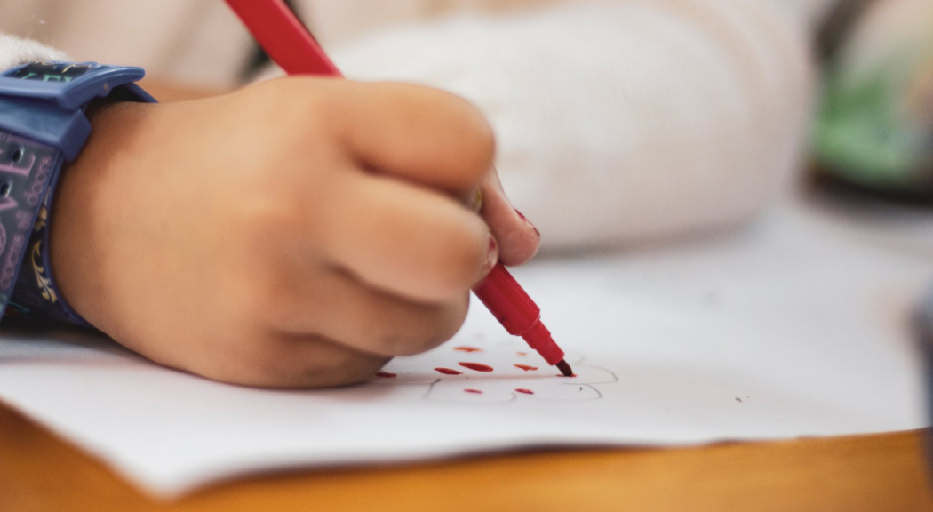 Primer plano de la mano de un niños escribiendo en un papel con un marcador rojo.