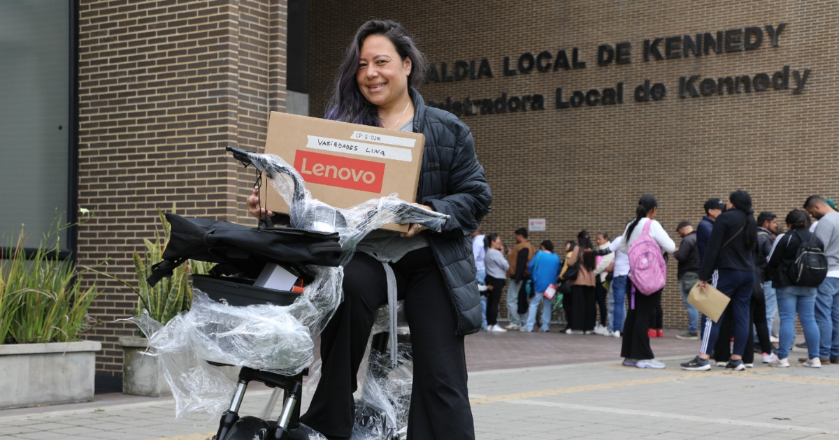 Foto de una mujer sonriente frente a la Alcaldía Local de Kennedy, sosteniendo una caja y una motocicleta eléctrica recién entregada.