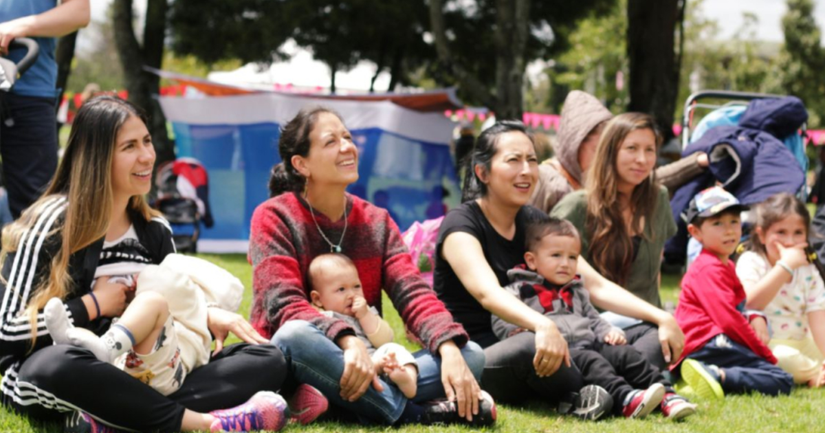 Imagen de mamás con niños en sus piernas sentadas en el pasto en un parque prestando atención a alguien, de fondo se ve la naturaleza del parque