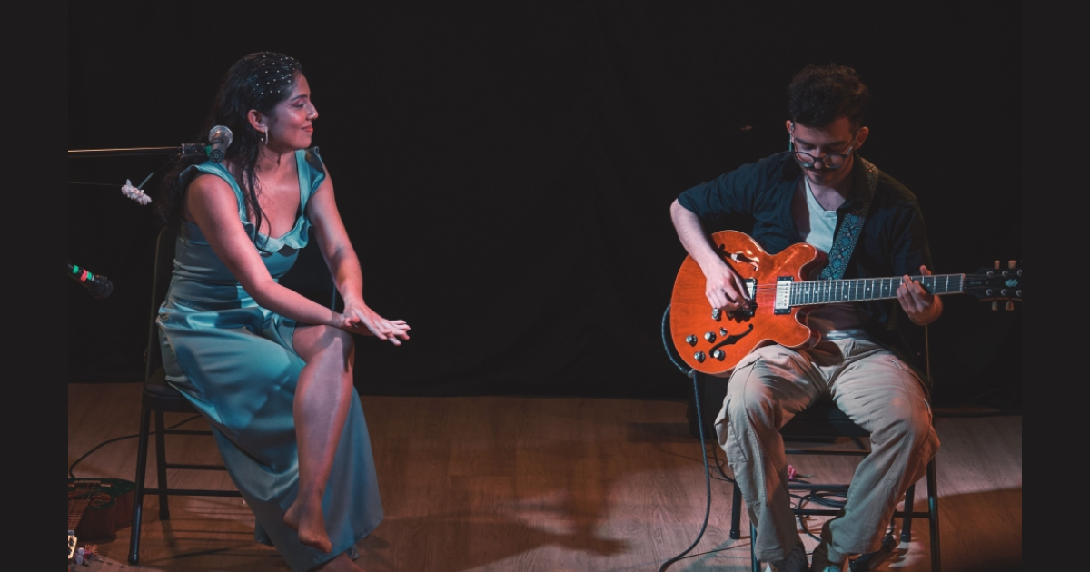 Imagen de una mujer sentada vestida de gala y de un hobre que toca guitarra en unescenario con fondo oscuro
