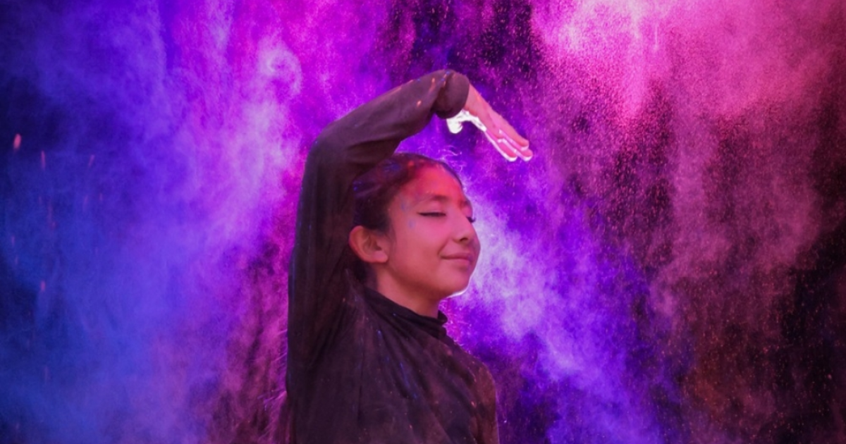 Imagen de una mujer jóven vestida de negro con su mano derecha en posición teatral, con un fondo de humo de colores