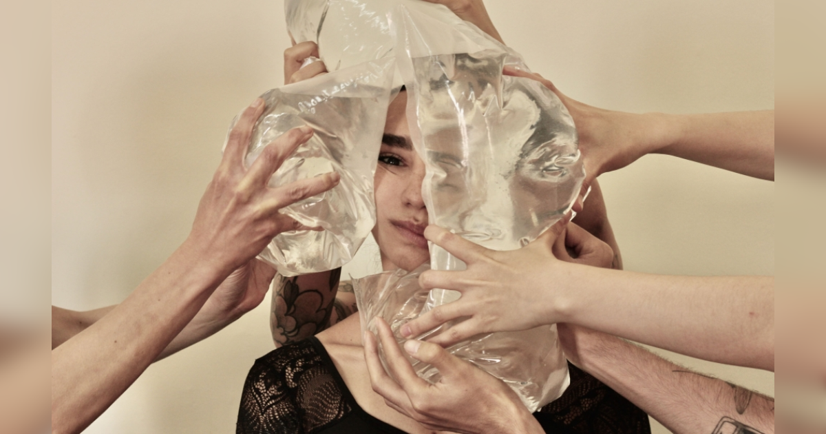 Imagen de la cara de una mujer, a su alrededor hay muchas manos de personas que tienen bolsas plasticas llenas de agua que al presionar su cara se ve desfigurada.