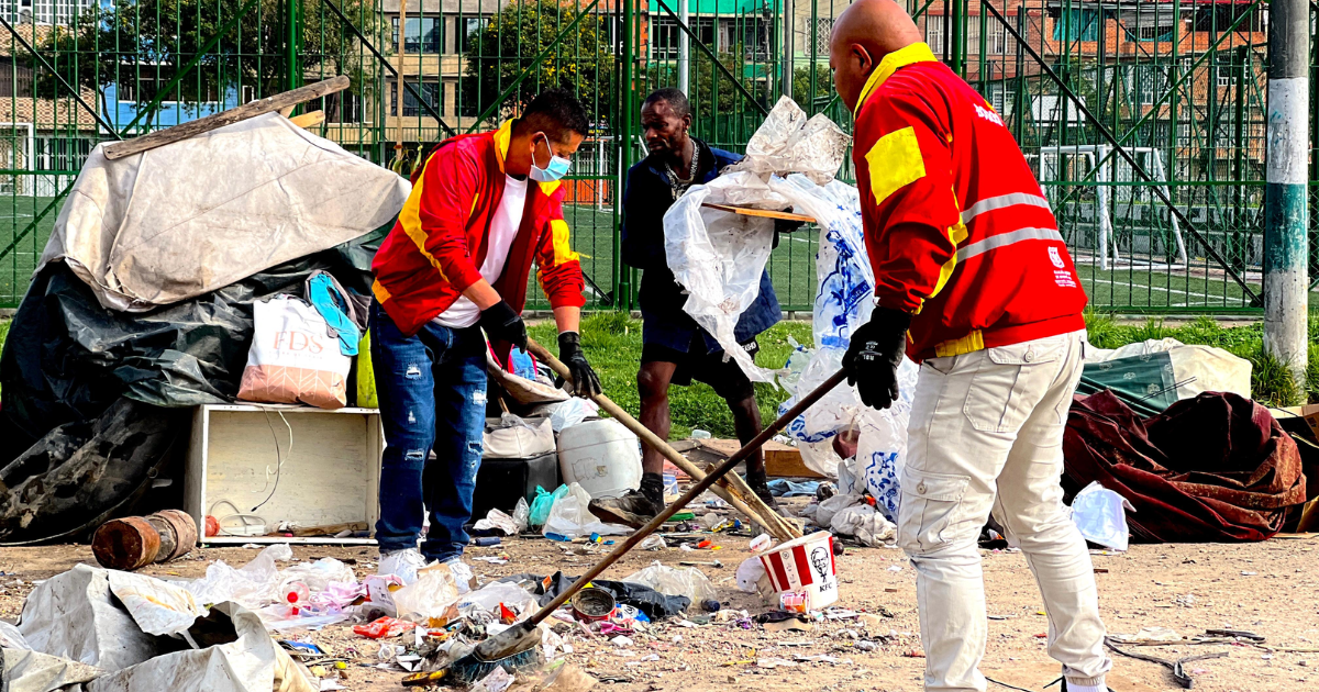 Foto de dos trabajadores de la Alcaldía Local de Kennedy, vestidos con chaquetas rojas, realizando labores de limpieza y recolección de basura en un espacio público