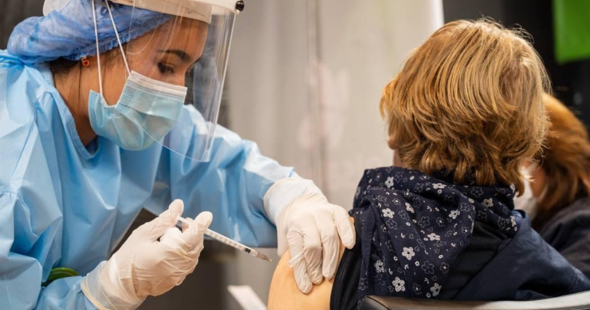 Imagen de una mujer con vestido se seguridad aplicando una vacuna a un mujer que está sentada en una silla