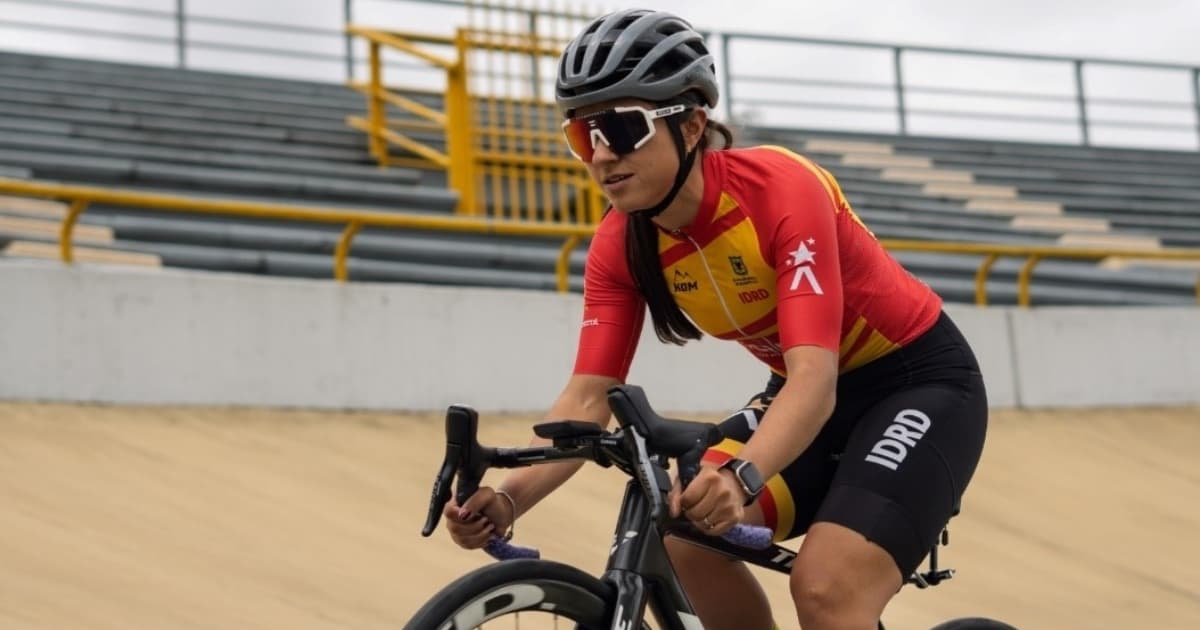 Paradeportista del Equipo Bogotá entrena ciclismo de pista en velódromo, usando casco y uniforme institucional rojo y amarillo, mientras pedalea sobre superficie inclinada con graderías metálicas al fondo.