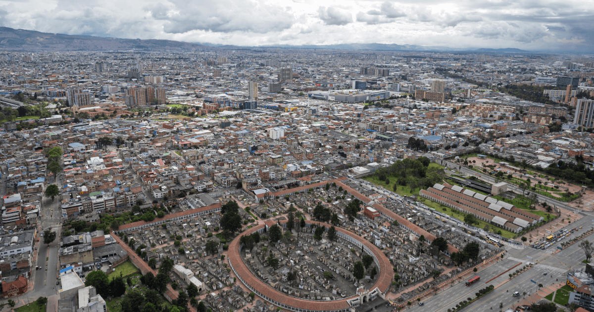 Vista aérea de Bogotá con extensa concentración de edificaciones, vías principales, cementerio central en primer plano y cielo cubierto de nubes sobre la ciudad.