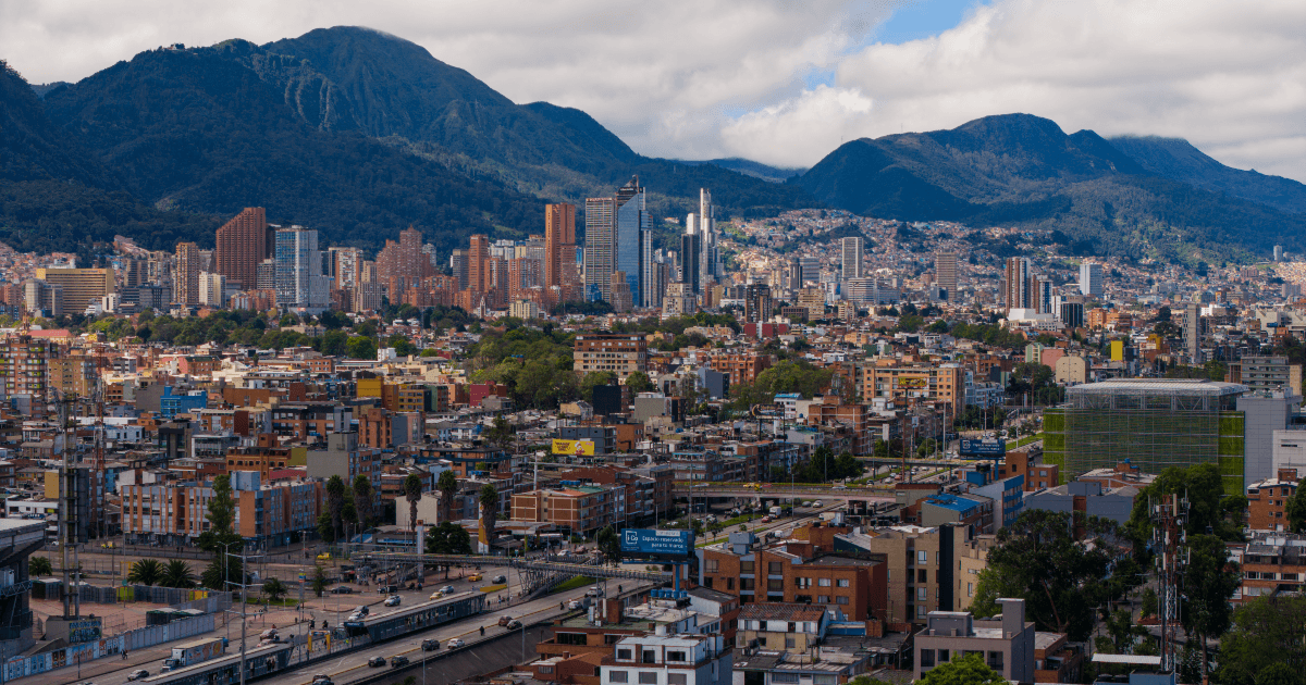 Vista panorámica de Bogotá con edificaciones residenciales y comerciales, vías principales y los cerros orientales al fondo bajo un cielo parcialmente nublado.