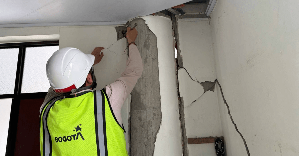 Persona con casco blanco y chaleco reflectivo con el logo de Bogotá inspecciona una columna y un muro interior con grietas visibles.