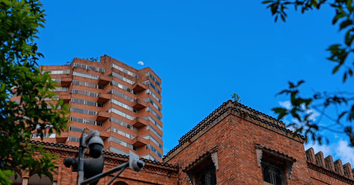 Edificaciones de ladrillo y torre de apartamentos en Bogotá, con vegetación en primer plano y cielo azul al fondo, en una escena urbana residencial.