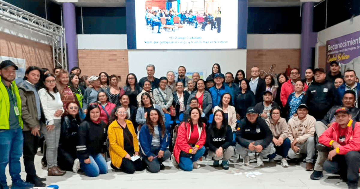 Grupo de personas participante en el Cuarto Diálogo Ciudadano del IDIGER en Ciudad Bolívar posan en un auditorio con una pantalla para proyectar presentaciones de fondo.