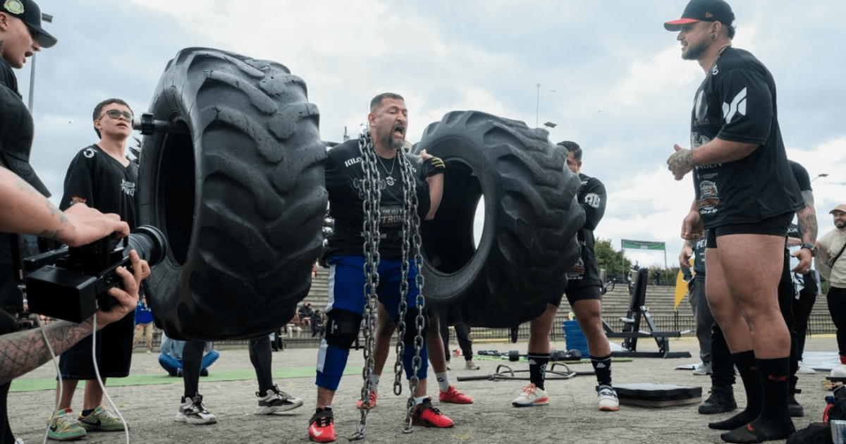 Persona participa en una prueba de fuerza al aire libre mientras levanta dos llantas de gran tamaño sujetas a una estructura metálica con cadenas.
