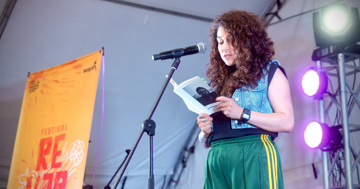 Imagen de una mujer joven en una tarima leyendo un libro de poesia, al lado inzquierdo se ve una pancarta del concurso Reverso.