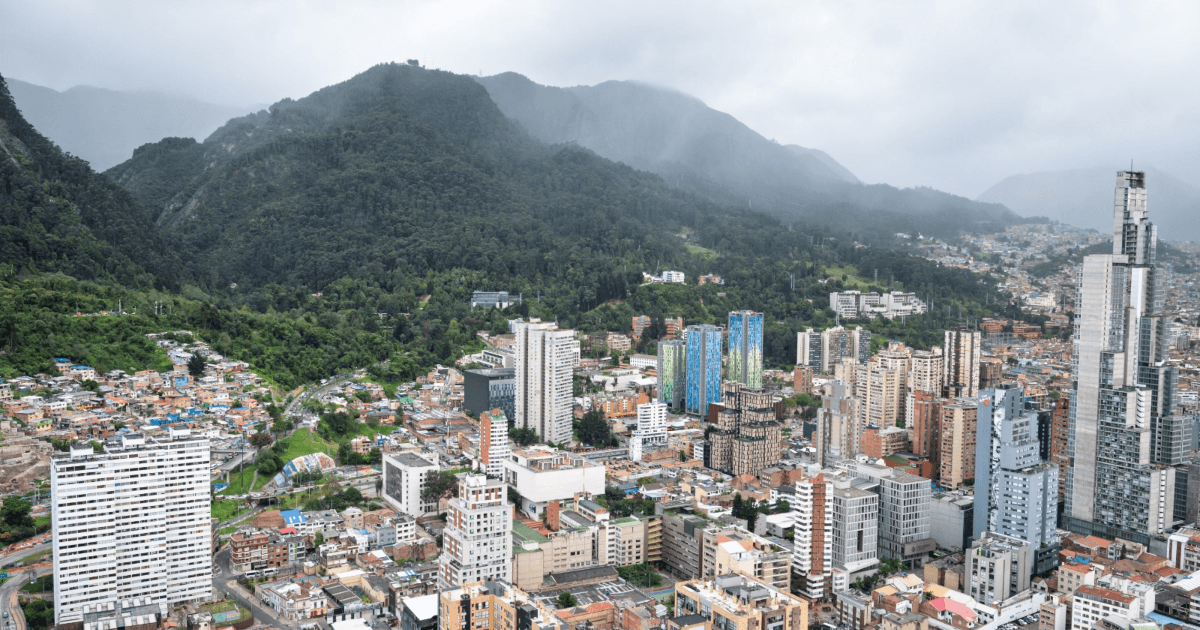 Vista panorámica de Bogotá con edificios residenciales y de oficinas en primer plano, zonas urbanas densamente construidas y los cerros orientales cubiertos de vegetación bajo un cielo nublado.