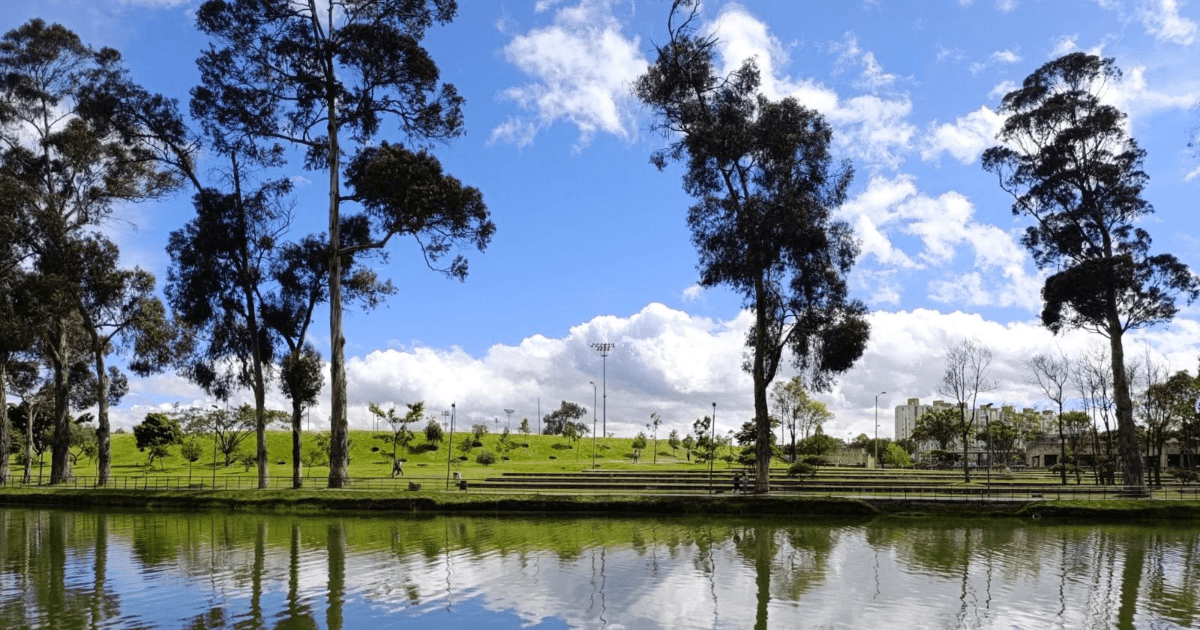 Cuerpo de agua que refleja el cielo y la vegetación circundante, rodeado por árboles altos y zonas de pasto verde. Al fondo se observan amplias áreas verdes y una fila de edificaciones bajo un cielo azul con nubes blancas dispersas.