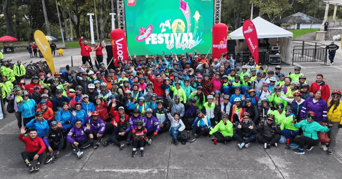 Fotografía grupal de cientos de ciclistas y asistentes al Festival de Verano, reunidos al aire libre frente a un escenario con una pantalla que anuncia la edición 29 del Festival de Verano.