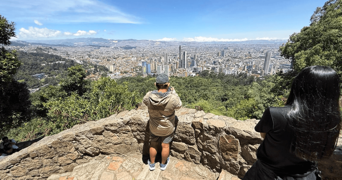 Vista panorámica de Bogotá desde un mirador en Monserrate. En primer plano se observa un muro de piedra y dos visitantes de espaldas contemplando la ciudad.