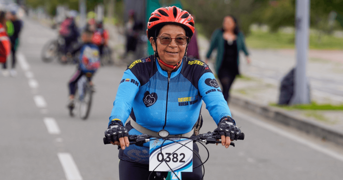 Participante de la Ciclovía de Bogotá se desplaza en bicicleta por una vía habilitada para la actividad. Lleva casco rojo, gafas y una camiseta deportiva azul con el número 0382 en la parte frontal.