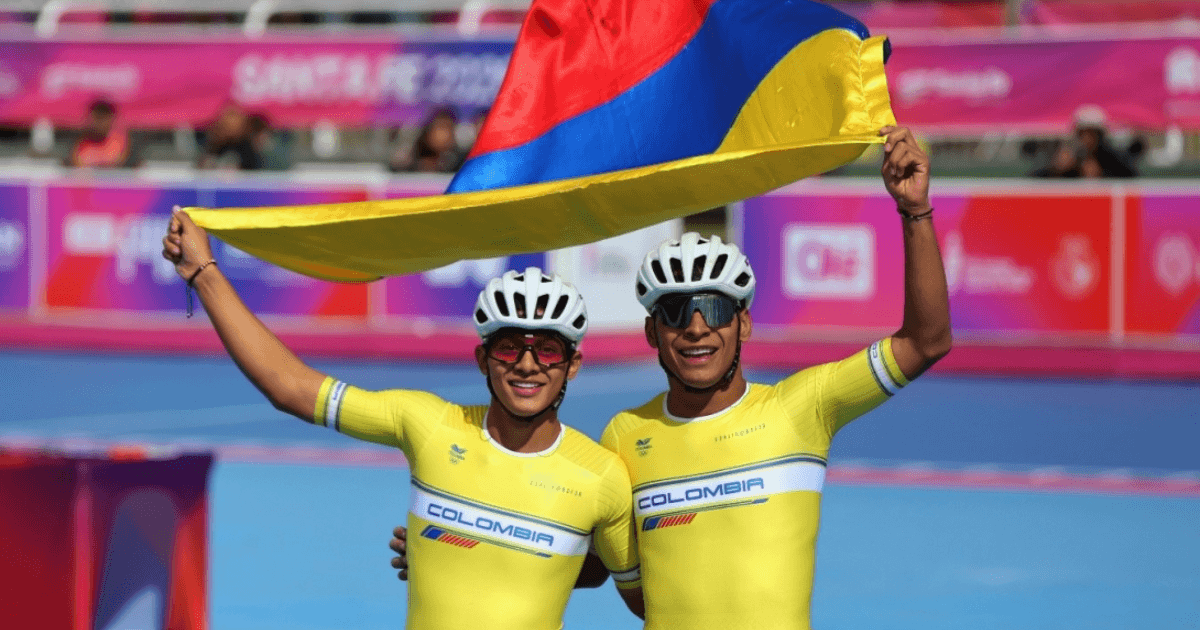 Dos deportistas de patinaje de velocidad con uniformes amarillos de Colombia y cascos blancos sostienen una bandera de Colombia sobre sus cabezas en una pista de competencia.
