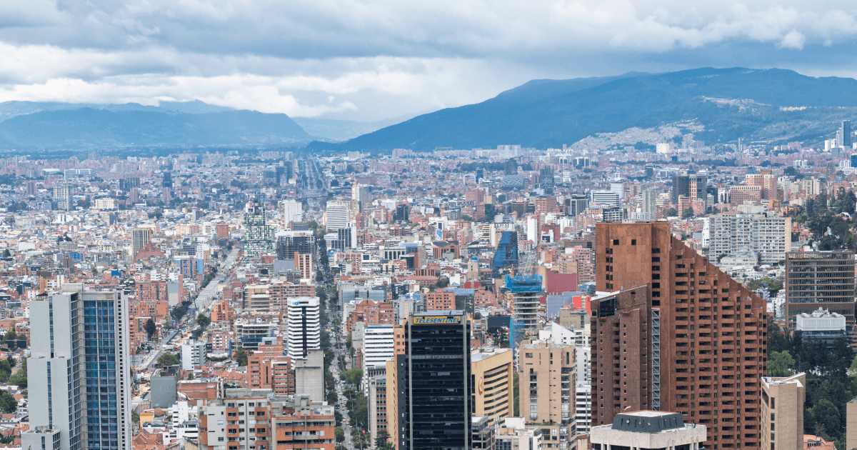 Vista panorámica de Bogotá con edificios residenciales y empresariales, avenidas principales y cerros al fondo bajo un cielo cubierto de nubes grises.