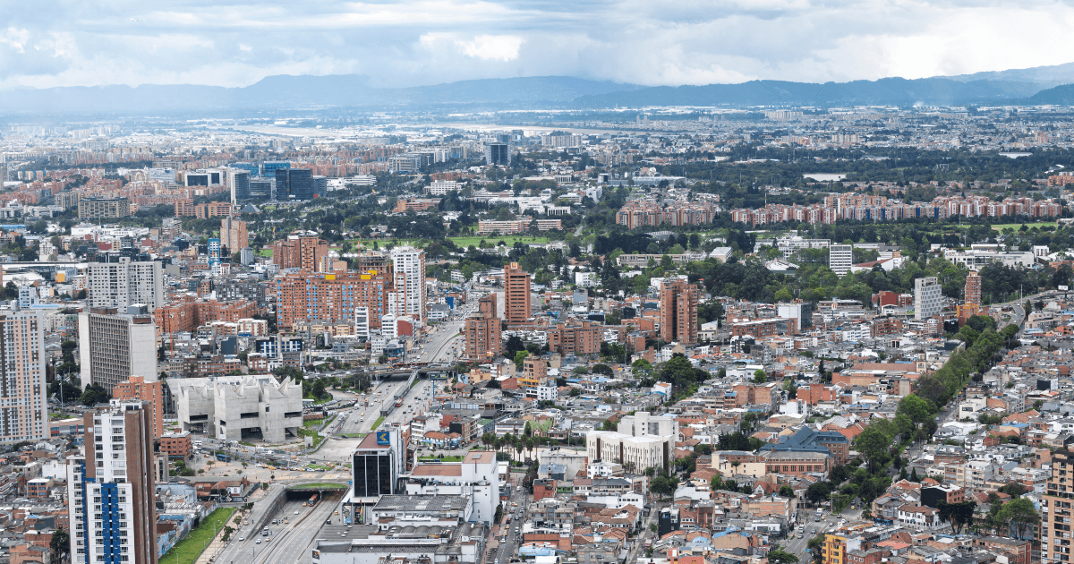 Vista panorámica de Bogotá con áreas residenciales, edificios de diferentes alturas, corredores viales y amplias zonas verdes. Al fondo se observan los cerros y un cielo cubierto de nubes sobre la ciudad.