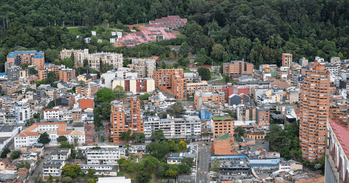 Vista panorámica de Bogotá con edificios residenciales y comerciales de diferentes alturas, calles arboladas y una extensa zona boscosa en los cerros orientales al fondo.