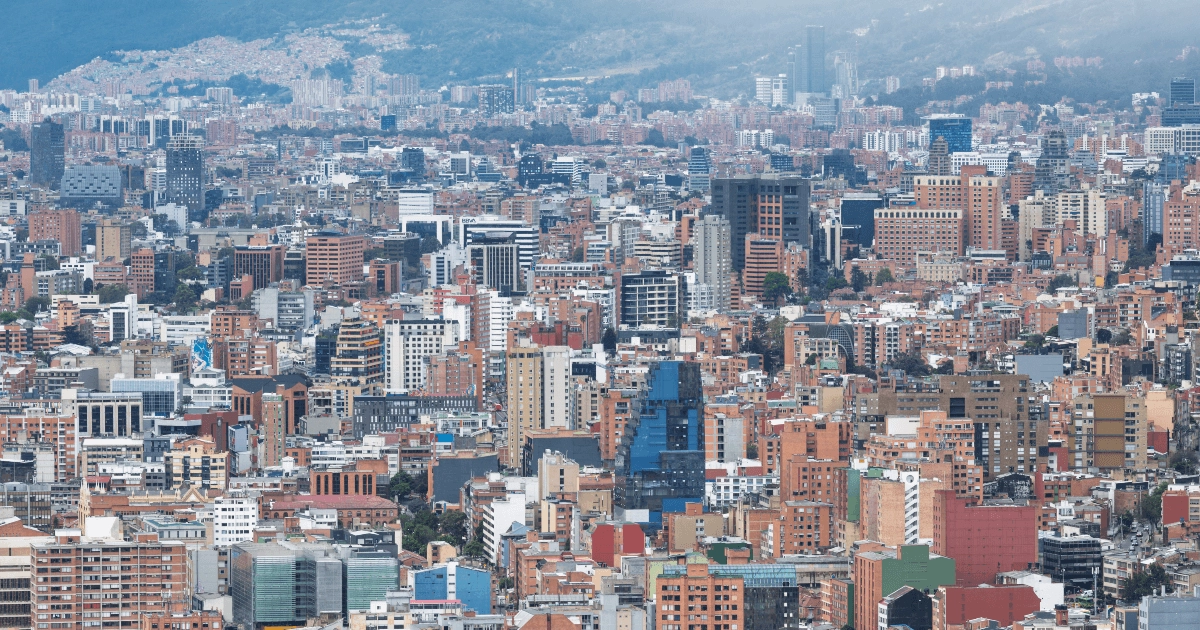 Vista panorámica del centro de Bogotá con numerosos edificios de diferentes alturas y colores, vías urbanas y montañas al fondo parcialmente cubiertas por neblina.