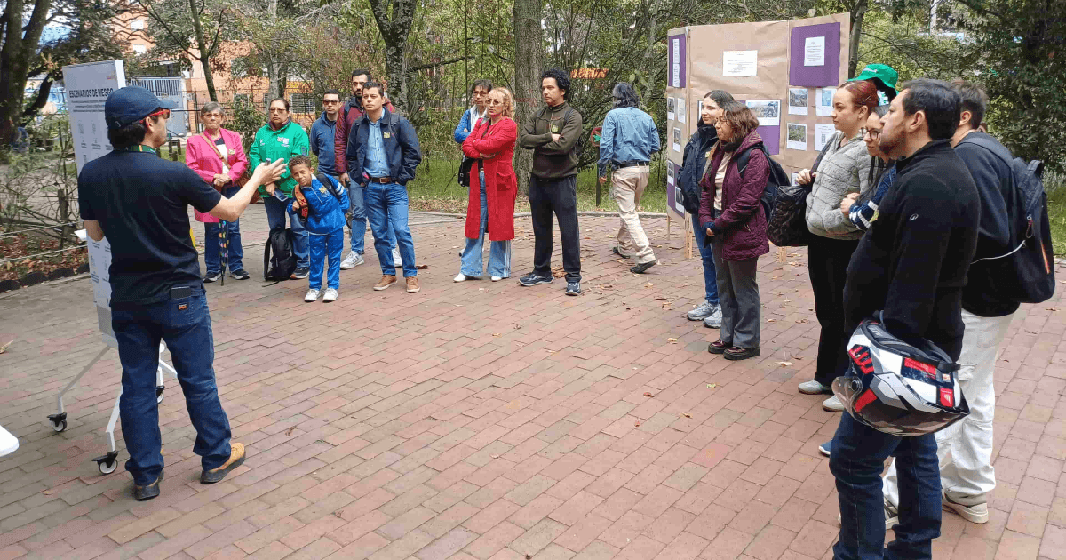 Personas participan en una actividad al aire libre sobre una plazoleta de ladrillo. Varias están alineadas y observan una demostración; algunas llevan chaquetas azules y una persona en el centro viste una chaqueta roja.
