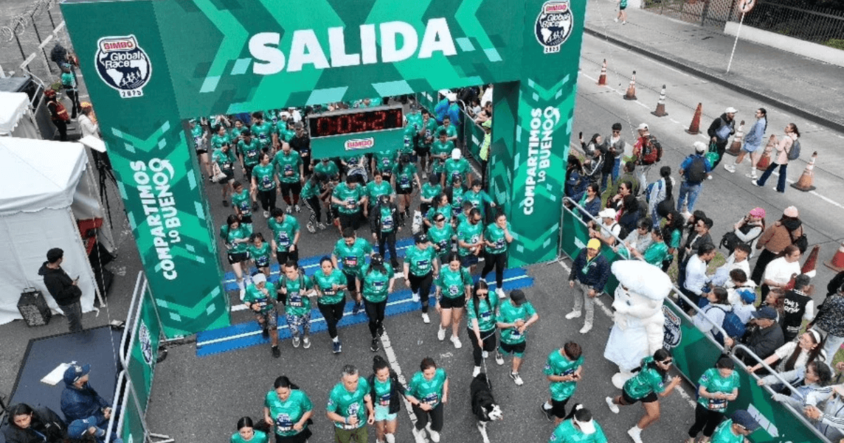 Cientos de participantes cruzan el arco de salida de la Bimbo Global Race 2026 sobre una vía urbana. El pórtico verde muestra el texto “SALIDA”, los logotipos de Bimbo Global Race 2025 y la frase “COMPARTIMOS LO BUENO”. A los lados hay espectadores detrás de vallas, mientras corredores con camisetas verdes avanzan por la línea de partida.