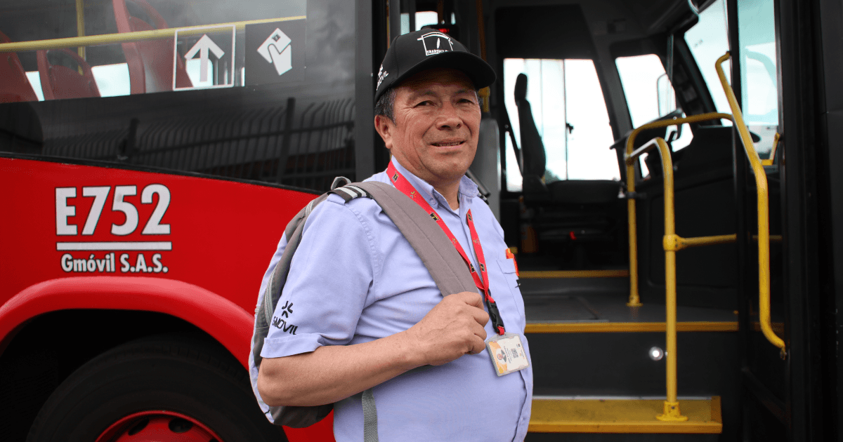 Persona con gorra negra, camisa azul claro y morral gris se encuentra junto a la puerta abierta de un bus rojo del sistema de transporte. En el costado del vehículo se observa la identificación “E752” y el nombre “Gmóvil S.A.S.”, mientras que el interior del autobús muestra pasamanos y escalones amarillos.