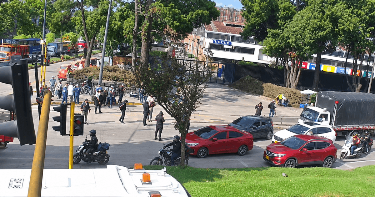 Vista general de una intersección vial en Bogotá con vehículos particulares, motocicletas y un camión detenidos. En un costado de la vía se observa un grupo de personas reunidas y presencia de operarios, mientras el tráfico avanza lentamente.