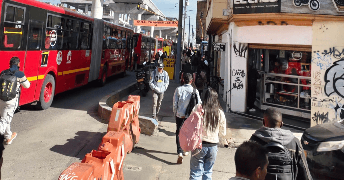 Peatones caminando por una acera junto a autobuses articulados rojos de TransMilenio en una concurrida vía urbana de Bogotá.