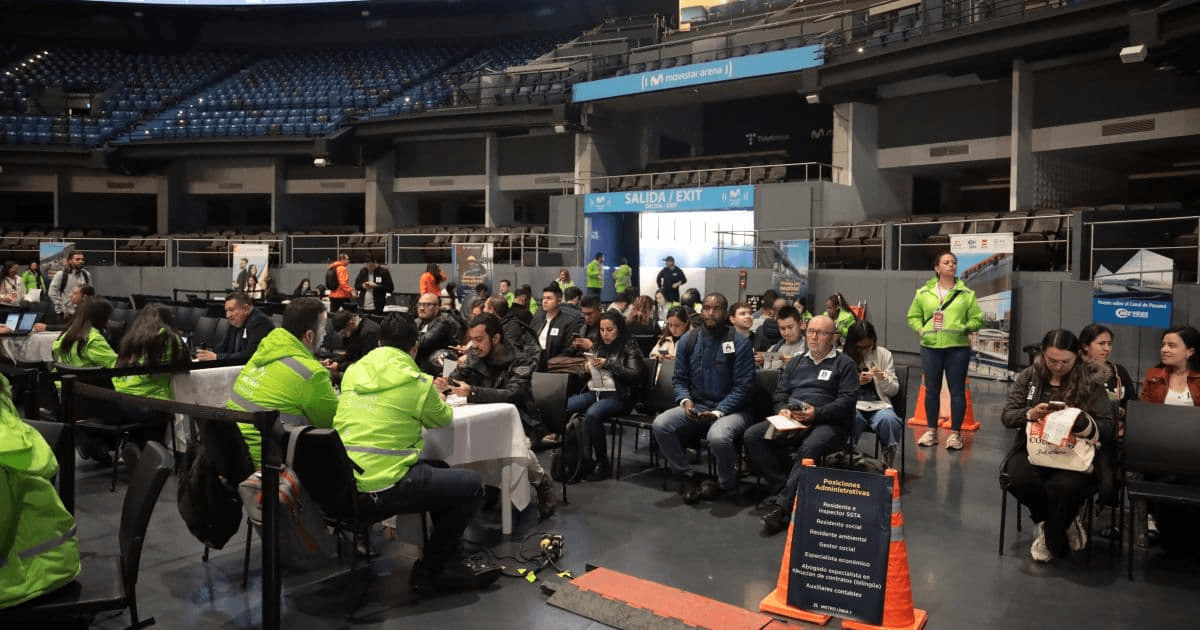 Personas sentadas en filas dentro de un recinto ferial mientras participan en una jornada de empleo. En primer plano hay mesas de atención y personas con chalecos institucionales; al fondo se observan estands, pantallas informativas y asistentes reunidos.
