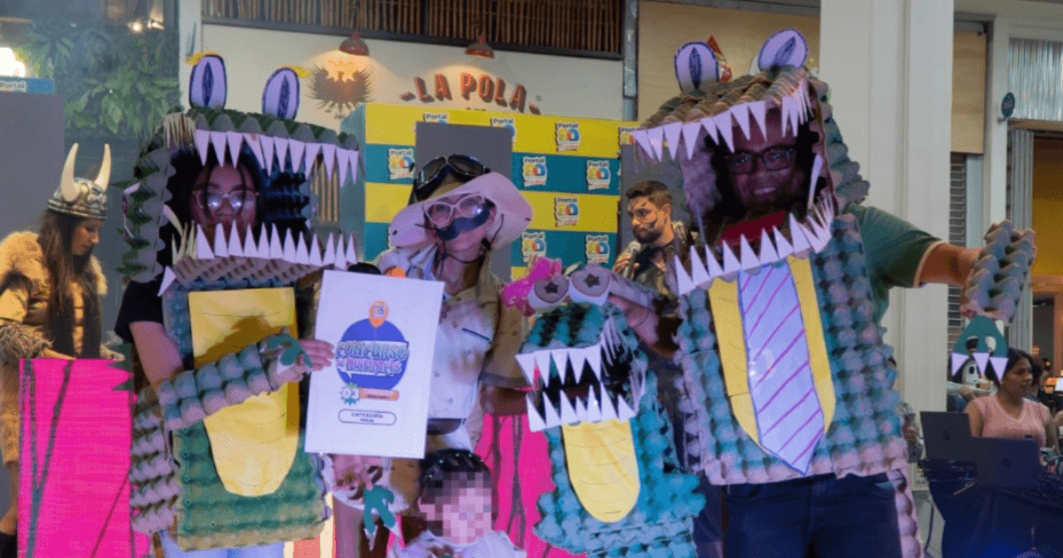 Grupo de personas posando en un centro comercial durante un concurso de disfraces. Dos personas llevan disfraces de cocodrilo hechos con cartones de huevera pintados de verde, con grandes mandíbulas abiertas, dientes blancos y ojos saltones.