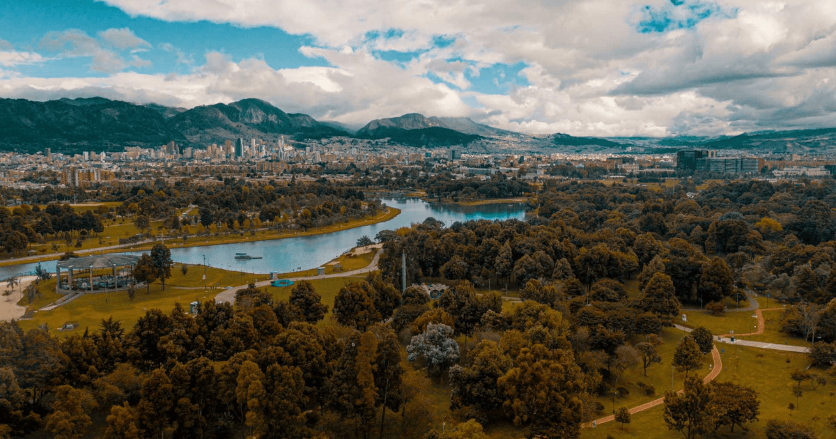 Vista aérea del parque Simón Bolívar en Bogotá, con zonas verdes, árboles, cuerpos de agua y los cerros orientales al fondo bajo un cielo parcialmente nublado.