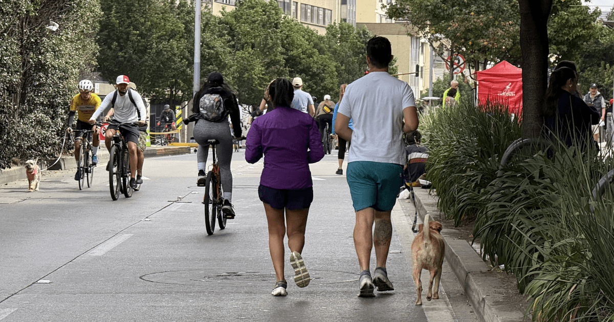 Personas caminando, corriendo y montando en bicicleta por una vía urbana cerrada al tráfico durante una jornada recreativa. Varias bicicletas circulan por la calzada y dos perros acompañan a los participantes en la Ciclovía de Bogotá.