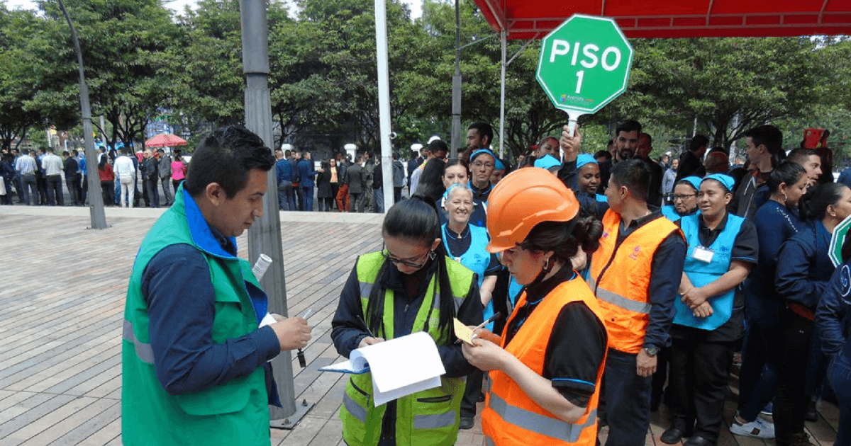 Personas participan en un ejercicio de evacuación en un espacio abierto. En primer plano, varios integrantes de brigadas de emergencia con chalecos reflectivos verdes y naranjas registran información en formularios. Detrás, un grupo organizado de participantes se reúne junto a una señal octagonal verde con el texto “PISO 1”.