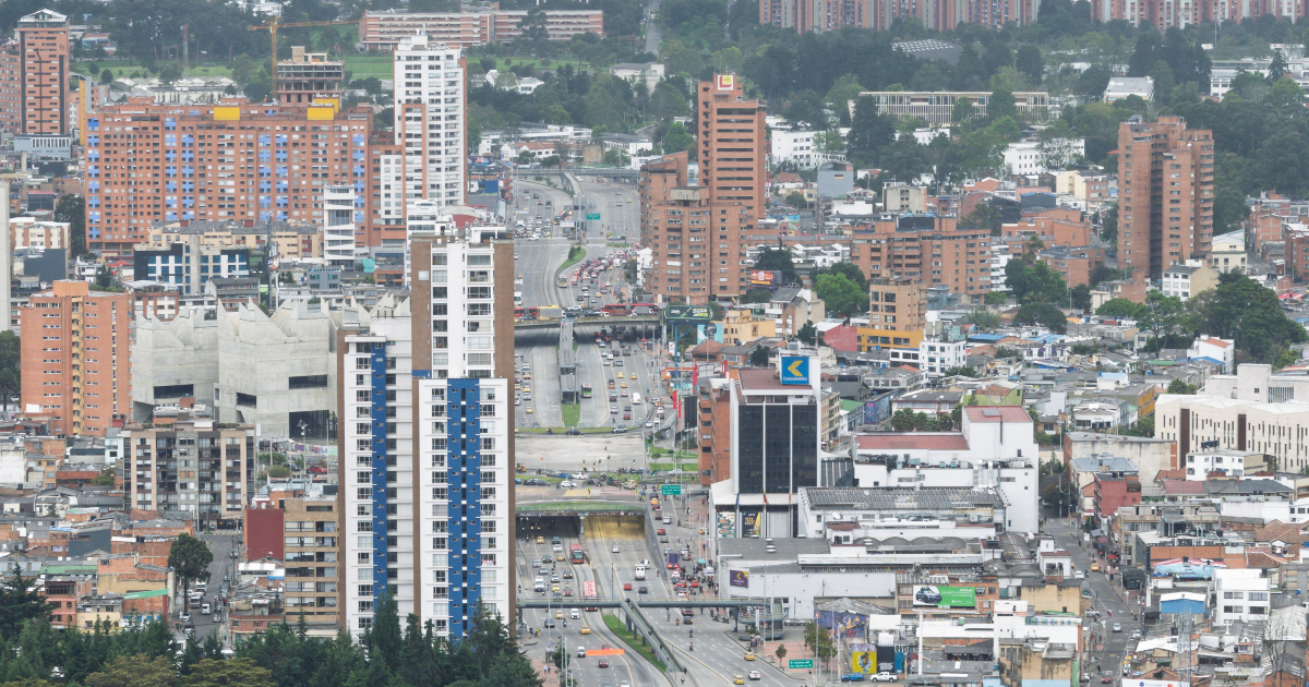 Vista panorámica de un sector urbano de Bogotá con edificios residenciales y comerciales de diferentes alturas, una vía principal con tráfico vehicular y construcciones densamente distribuidas en el paisaje urbano.