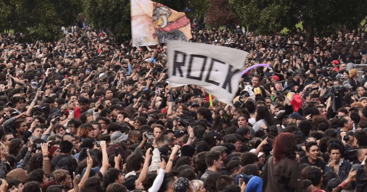Multitud de asistentes reunidos al aire libre durante Rock al Parque en Bogotá. En el centro se observa una bandera blanca con la palabra “ROCK”, mientras cientos de personas ocupan gran parte del espacio frente al escenario, rodeadas por árboles.