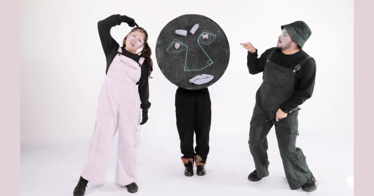 Imagen de tres personajes, uno viste un overol rosado y el otro verde oscuro, el tercer personaje lleva un gran círculo negro con un rostro dibujado en tiza que cubre su cuerpo.