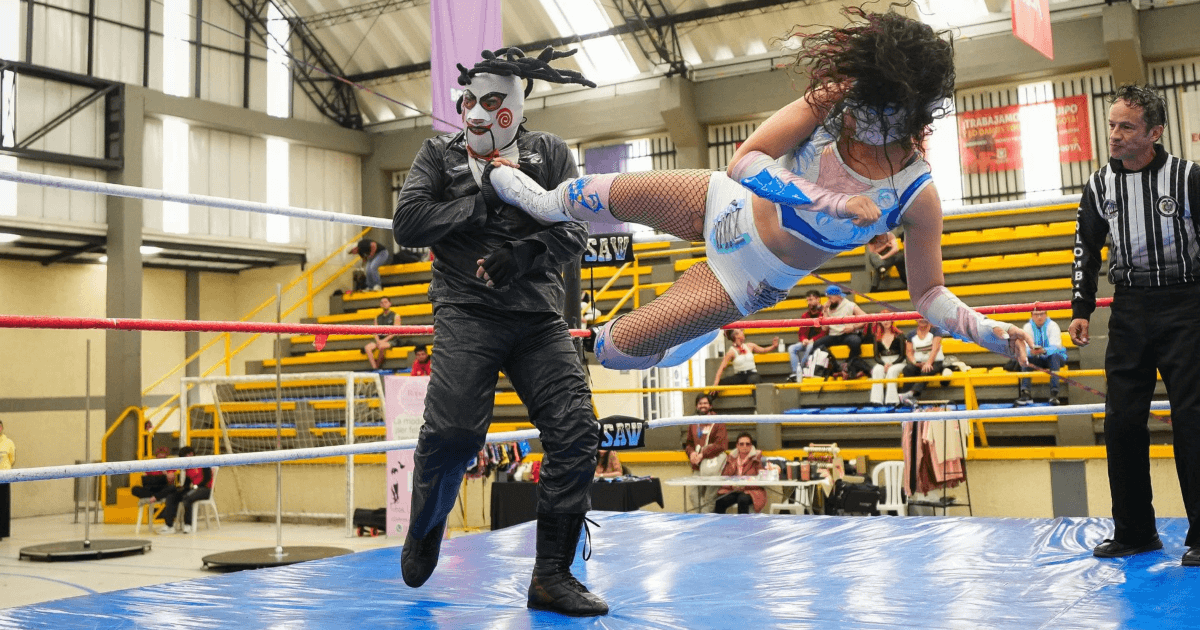 Una competidora de lucha libre realiza una patada aérea contra un luchador enmascarado dentro del cuadrilátero, mientras un árbitro y espectadores observan el combate.