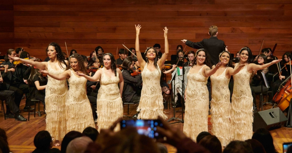 Imagen de varias mujeres vestidas con trajes de danza oriental. De fondo se ve una agrupación musical.