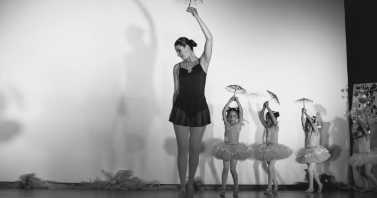 Imagen de una mujer vestida de traje negro de ballet y cuatro niñas vestidas de ballet con una pequeña sombrilla en sus manos.