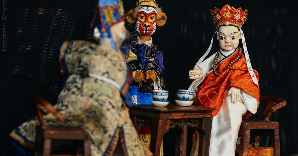 Imagen de tres títeres de tradición oriental representando una escena teatral alrededor de una mesa con tazas, sobre un fondo oscuro.
