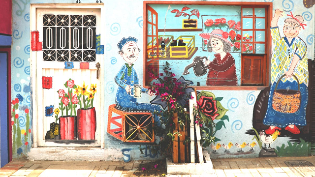 La pared de una casa pintada con dos personas mayores regando las plantas, hay una buganvilia creciendo al lado de la puerta