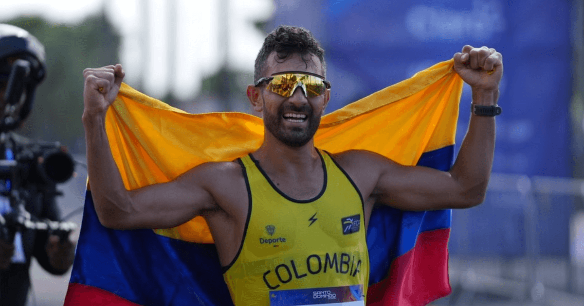 Eider Arévalo con uniforme amarillo de Colombia y gafas deportivas sostiene la bandera nacional extendida detrás de la espalda durante una competencia de marcha atlética.