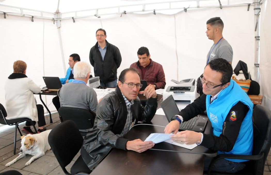 Un hombre en chaqueta azul atiende a un ciudadano en una mesa, detrás de ellos hay varias personas trabajando en conjunto 