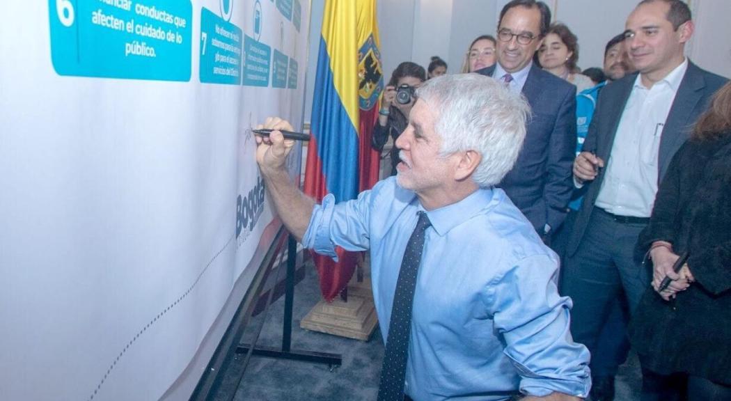 Alcalde Peñalosa viste camisa y corbata azul y se agacha para firmar un tablero.
