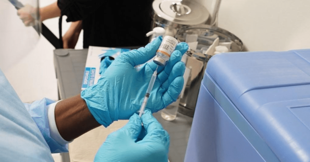 Imagen de unas manos que usan guantes y tienen una geringa y frasco de dosis de vacuna