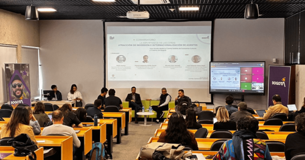 Imagen de salón donde tres personas exponen cifras con una gran imagen con los resultados, de frente un auditorio presta atención