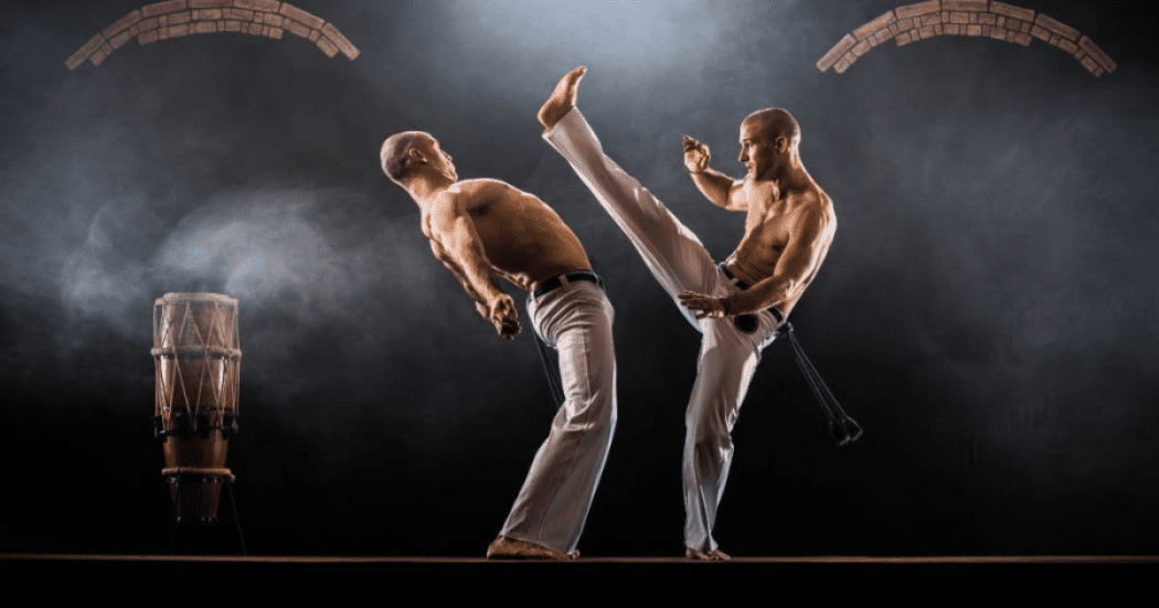 Imagen de dos hombres de pantalon blanco y sin camisa practicando capoeira